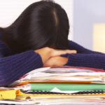 woman with long, dark hair with head down on top of a pile of folders and books