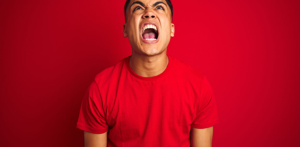Young brazilian man wearing t-shirt standing over isolated red background angry and mad screaming frustrated and furious, shouting with anger. Rage and aggressive concept.