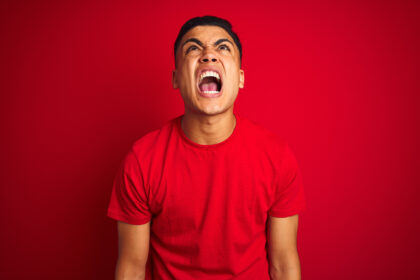 Young brazilian man wearing t-shirt standing over isolated red background angry and mad screaming frustrated and furious, shouting with anger. Rage and aggressive concept.