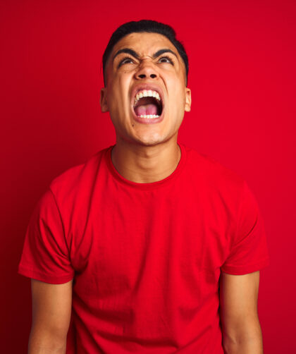 Young brazilian man wearing t-shirt standing over isolated red background angry and mad screaming frustrated and furious, shouting with anger. Rage and aggressive concept.