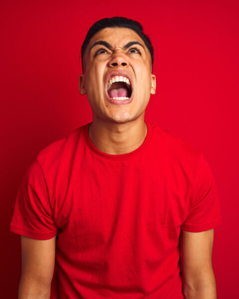 Young brazilian man wearing t-shirt standing over isolated red background angry and mad screaming frustrated and furious, shouting with anger. Rage and aggressive concept.