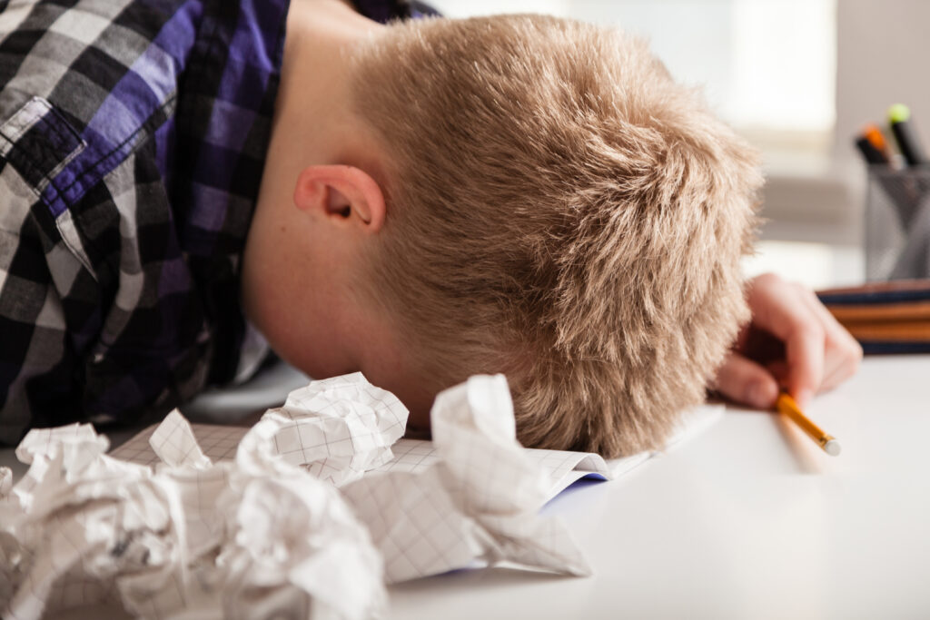 Young boy struggling with his homework sitting with his head down on the table surrounded by crumpled pages of screwed up paper