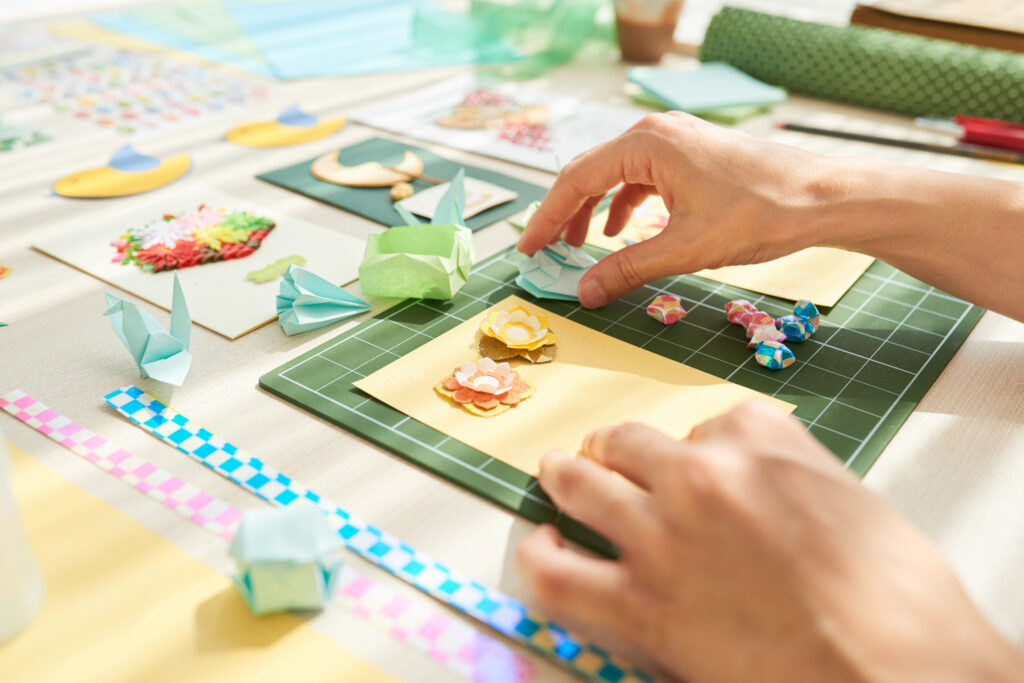 Creation of unique handmade greeting cards: unrecognizable woman sitting at wooden table and gluing paper flowers on cardboard, close-up shot