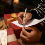 Pen, hands and man with card and gift for Christmas event or party at home for family. Celebration, paper and closeup of male person writing letter with present boxes for xmas festive holiday