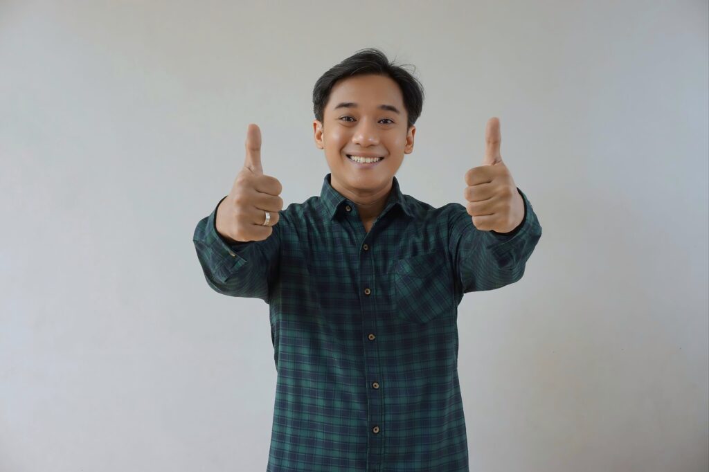 Adult Asian man in green long sleeve flannel shirt looking at camera while smiling and showing two thumbs up. Happy and cheerful expression. Gesture of achievement, appreciation, agreement, approval.