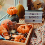 Autumn cozy mood composition on the windowsill. Pumpkins, cones, candles on wooden tray, blurred Fall mood message on lightbox, warm plaid. Autumn, fall, hygge home decor. Selective focus. Copy space